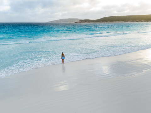 Aerial view of a woman standing on the white sand of Hellfire Bay with turquoise waves gently lapping the shore in Cape Le Grand National Park, Western Australia, Australia.