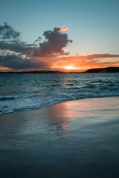 View of Muttonbird Beach at sunset with gentle waves washing onto the wet sand, reflecting the orange and pink hues of the sky in Elleker, Western Australia, Australia.