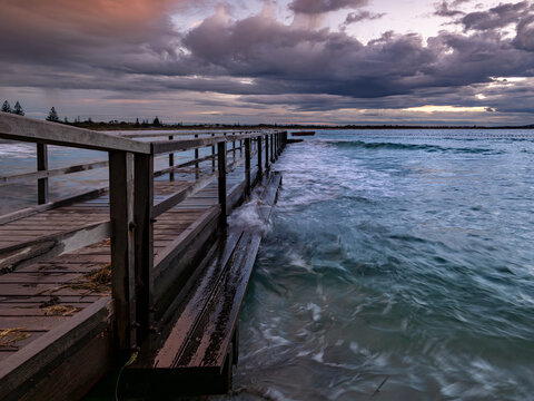 View of the wooden jetty with gentle waves lapping against the structure under a dramatic cloudy sky at dusk, Emu Point, Western Australia, Australia.