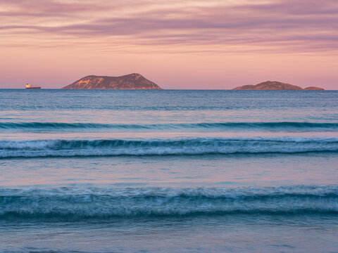 View of gentle ocean waves rolling onto the shore with distant islands and a cargo ship on the horizon under a soft pink sunset sky at Middleton Beach, Western Australia, Australia.