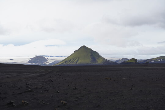 View of the moss-covered Maelifell volcano rising from the black volcanic sand plains with a distant glacier under a cloudy sky in Kirkjubaejarklaustur, Skaftarhreppur, Iceland.