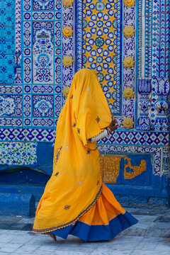 View of a woman wearing a vibrant yellow and blue traditional outfit walking past the intricately tiled walls of the Shrine of Sachal Sarmast in Daraza Sharif, Sindh, Pakistan.