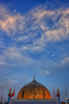 View of the golden dome of the Shrine of Lal Shahbaz Qalandar adorned with flags under a clear blue sky with wispy clouds and a crescent moon in Sehwan, Sindh, Pakistan.
