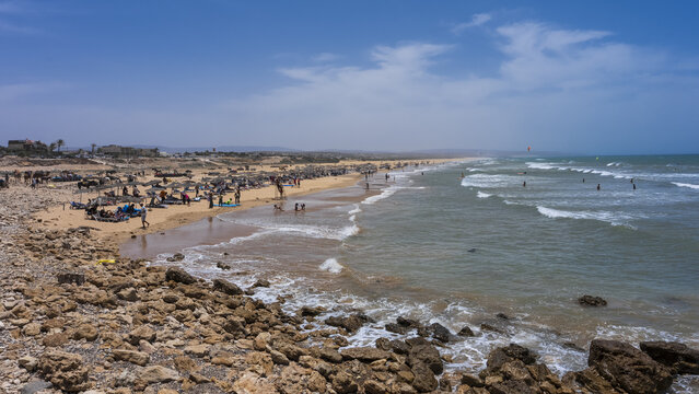 View of the sandy beach at Sidi Kaouki with a rocky foreground, people relaxing under umbrellas, and gentle ocean waves under a clear blue sky in Sidi Kaouki, Marrakesh-Safi, Morocco.