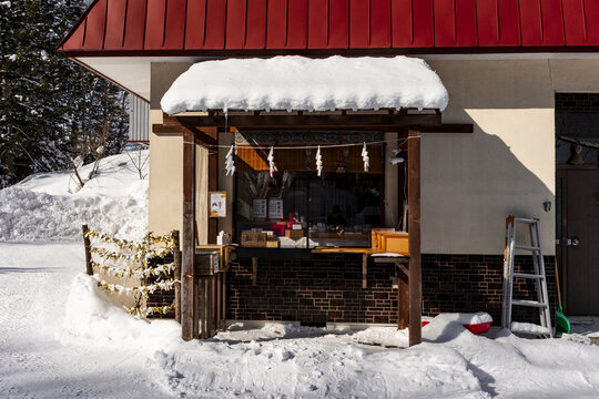 View of a snow-covered wooden shrine stall with shimenawa rope and paper charms in a winter landscape in Niseko, Hokkaido, Japan.