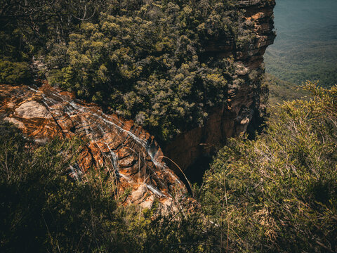 View of the Blue Mountains featuring cascading waterfalls flowing over rugged sandstone cliffs surrounded by dense eucalyptus forest in The Blue Mountains, New South Wales, Australia.