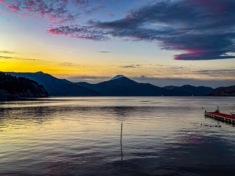 View of Lake Ashinoko at sunset with Mount Fuji visible in the distance, calm water reflecting the colorful sky, and a wooden pier in Hakone, Kanagawa, Japan.
