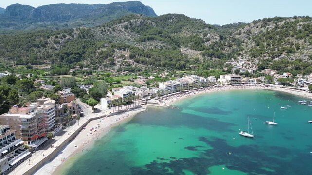 Aerial view of the turquoise bay and beach of Port de Soller with boats anchored in the water, surrounded by coastal hotels and mountains in Port de Soller, Balearic Islands, Spain.