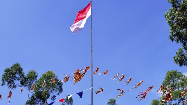 Indonesian flag waving on pole with red-white colors against blue sky and green trees concept representing national symbol, patriotism, independence, sovereignty, and democratic identity.