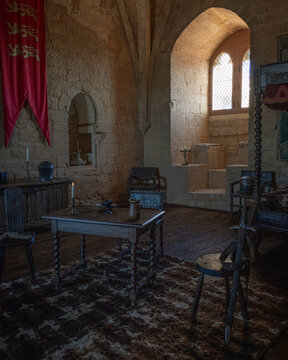 View of the interior of a medieval room at the Chateau de Beynac featuring stone walls, a wooden table, rustic chairs, and a red banner in Beynac-et-Cazenac, Nouvelle-Aquitaine, France.