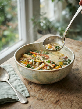 Vegetable soup being ladled from bowl with steam rising by window