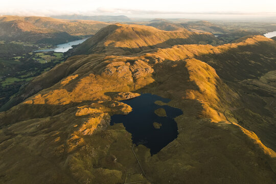 Aerial view of Angletarn Pikes featuring the dark waters of Angle Tarn surrounded by rolling grassy hills and rocky terrain under golden hour light in Penrith, England, United Kingdom.