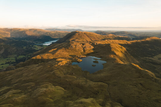 Aerial view of Angletarn Pikes and Angle Tarn surrounded by rolling hills and grassy terrain under golden hour light in Penrith, England, United Kingdom.