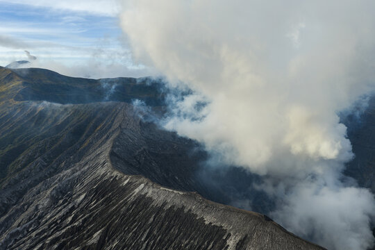 Aerial view of Mount Bromo crater with thick volcanic smoke rising from the rugged landscape in Tosari, East Java, Indonesia.
