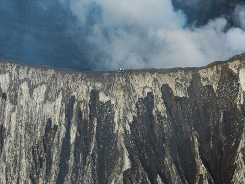 Aerial view of the Mount Bromo crater rim with people standing on the edge, volcanic ash, and sulfurous smoke rising from the steep textured slopes in Tosari, East Java, Indonesia.