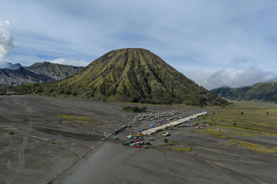 Aerial view of Mount Bromo with its iconic volcanic cone rising above the vast volcanic sand sea, featuring parked jeeps and small structures at the base in Tosari, East Java, Indonesia.