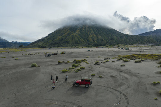 Aerial view of Mount Bromo with a red off-road vehicle parked on the sandy volcanic plain and people standing nearby in Tosari, East Java, Indonesia.