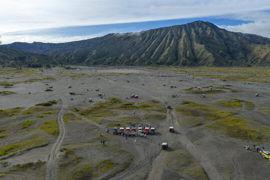 Aerial view of Mount Bromo and the vast volcanic sand sea with off-road vehicles and tourists gathered in the foreground in Tosari, East Java, Indonesia.