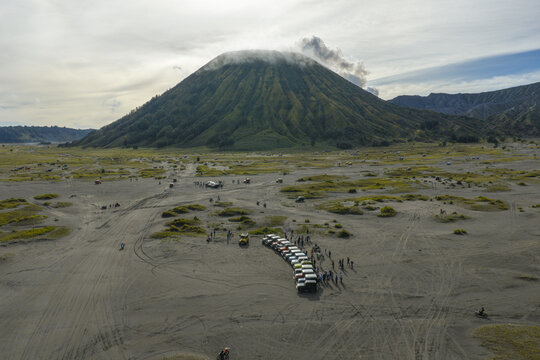 Aerial view of Mount Bromo with a line of parked jeeps on the sandy volcanic plain under a cloudy sky in Tosari, East Java, Indonesia.