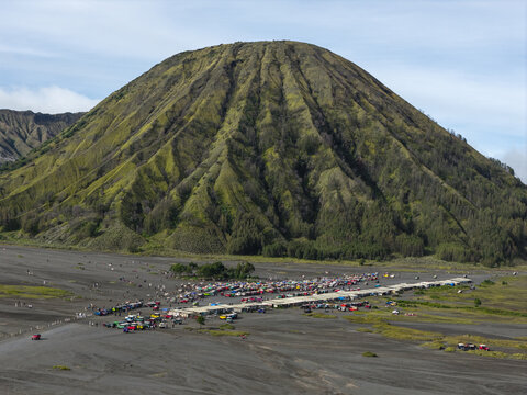 Aerial view of Mount Bromo volcano with its distinctive ridged slopes and a large group of parked off-road vehicles in the sandy caldera below in Tosari, East Java, Indonesia.