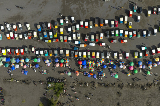 Aerial view of Mount Bromo with numerous off-road jeeps parked in rows and colorful umbrellas scattered across the volcanic sand in Tosari, East Java, Indonesia.