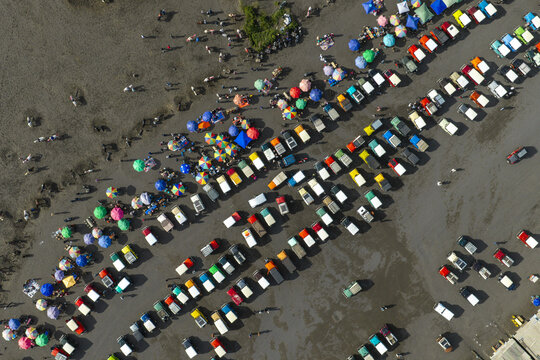 Aerial view of numerous colorful off-road jeeps parked in a large sandy area near Mount Bromo, with people walking between vehicles in Tosari, East Java, Indonesia.