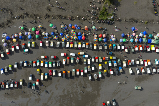 Aerial view of many colorful off-road jeeps parked in rows on the volcanic sand near Mount Bromo, with vibrant umbrellas and people walking between vehicles in Tosari, East Java, Indonesia.