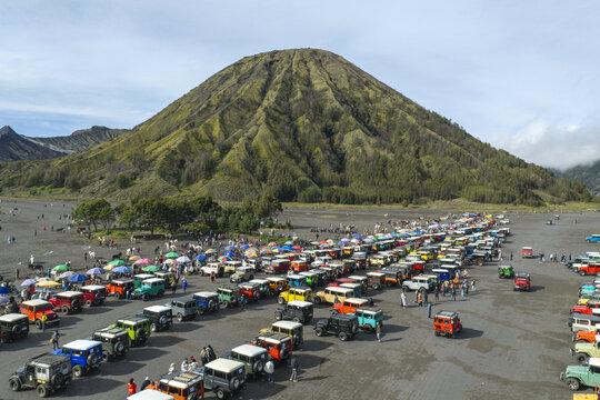 Aerial view of Mount Bromo rising above a vast volcanic sand plain filled with numerous colorful 4x4 off-road vehicles and tourists in Tosari, East Java, Indonesia.