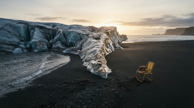 Wooden chair on a black sand beach beside a glacier under a cloudy sunset