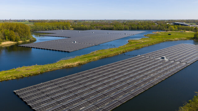 Aerial view of a large floating solar farm with rows of photovoltaic panels installed on a calm lake surrounded by green banks and trees in Zwolle, Overijssel, Netherlands.