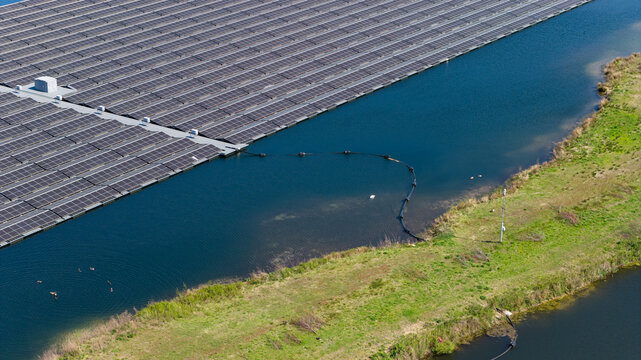 Aerial view of a large floating solar farm installed on a calm lake, with green grassy banks and ducks swimming in the water in Zwolle, Overijssel, Netherlands.