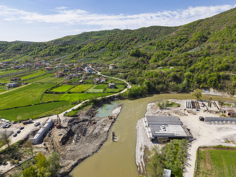 Aerial view of bridge construction site over a river with concrete beams, construction equipment, and surrounding rural landscape in Paper, Elbasan County, Albania.