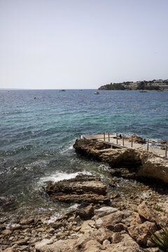 View of a wooden pier extending over the rocky coastline into the turquoise Mediterranean sea in Palma Nova, Balearic Islands, Spain.