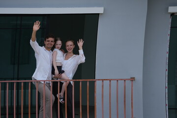 Obraz premium Caucasian family of three embracing wave happily from their balcony, the girl riding piggyback on the balcony of their new house overlooking the residential estate, authentic everyday scene