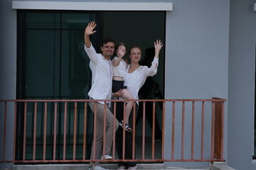 Obraz premium Caucasian family of three embracing laugh and wave from the new balcony railing standing on the terrace of a newly built suburban home. Healthy active lifestyle of a modern family