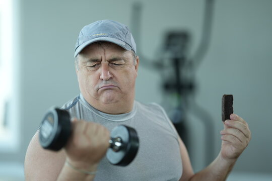 Funny fat senior man on a diet weighs iron against chocolate in a comic dilemma while trying to start a healthy diet in the gym. Getting in shape is hard, humorous fitness moment