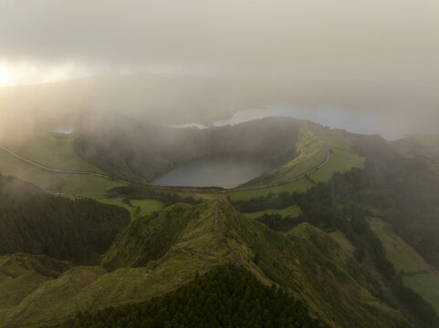Aerial view of Sete Cidades volcanic crater lakes surrounded by lush green hills and winding roads under a misty sky in Sete Cidades, Azores, Portugal.