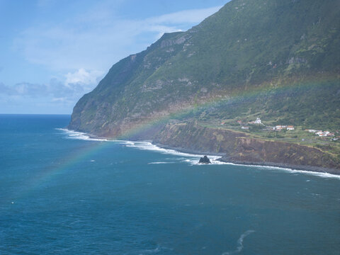 Aerial view of Faja Grande with a vibrant rainbow arching over the dramatic coastal cliffs, lush green hills, and the deep blue Atlantic Ocean in Faja Grande, Azores, Portugal.