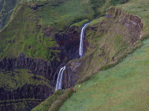 Aerial view of Cascata da Ribeira Grande cascading down lush green cliffs with two hikers standing on the grassy edge in Faja Grande, Acores, Portugal.