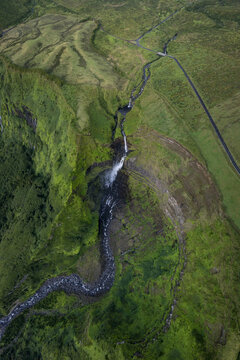 Aerial view of Cascata da Ribeira Grande cascading down lush green cliffs into a winding stream with a road visible in the landscape in Faja Grande, Acores, Portugal.
