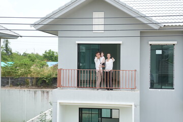Obraz premium Happy young family with little daughter stand at the wooden railing admiring the neighborhood view on the upper balcony of their new home above the neighborhood, authentic everyday scene