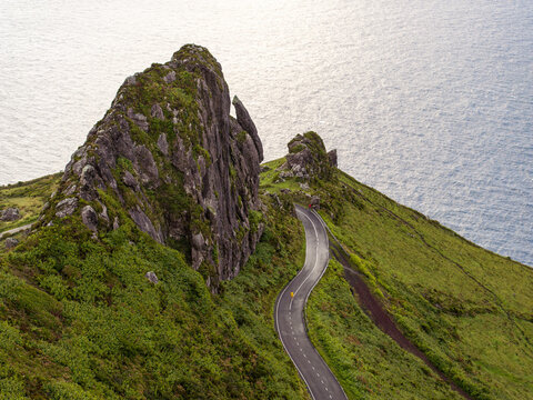 Aerial view of Rocha dos Frades with a winding coastal road cutting through lush green hills overlooking the Atlantic Ocean in Lajes das Flores, Azores, Portugal.
