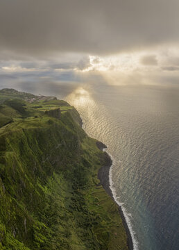 Aerial view of Rocha dos Frades with dramatic green cliffs, a black sand beach, and sun rays breaking through clouds over the Atlantic Ocean in Lajes das Flores, Azores, Portugal.