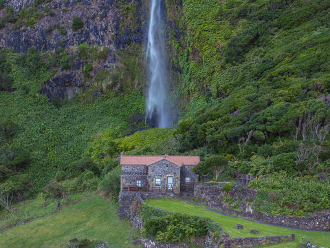 Aerial view of Moinho da Cascata nestled at the base of a towering waterfall surrounded by lush green cliffs and stone walls in Faja Grande, Acores, Portugal.