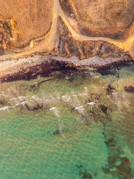 Aerial view of the rugged coastline of Kos with golden cliffs meeting the clear turquoise waters of the Aegean Sea under warm sunlight, Kos, Greece.