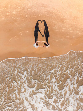 Aerial view of two people on a sandy beach forming a heart shape with their hands and shadows near the waves in Kos, Greece.