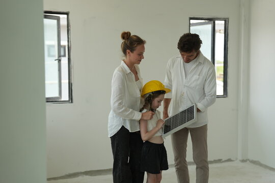 Young parents teaching their curious child read the specification sheet while the girl wears a yellow helmet inside their new energy efficient home in warm natural light