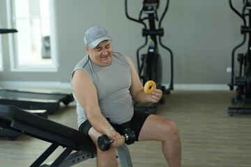 Obraz premium Overweight middle aged man in gray cap sits on the incline bench holding a dumbbell and a tempting donut during his weight loss workout at the fitness club in warm natural light