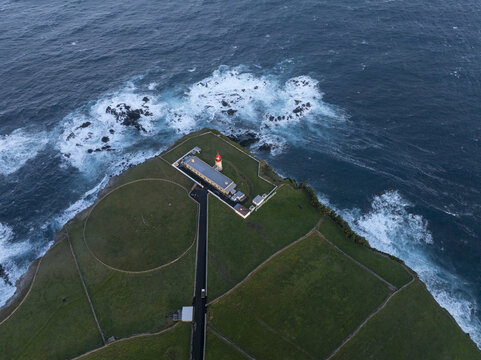 Aerial view of Farol de Alvernaz lighthouse perched on a green cliff edge overlooking the deep blue Atlantic Ocean with crashing waves and stone walls in Ponta Delgada, Acores, Portugal.