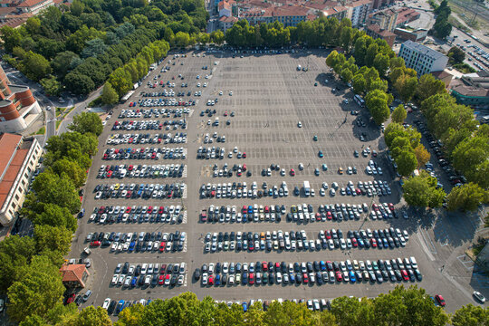 Aerial view of a large parking lot filled with many cars, surrounded by lush green trees and urban buildings in Asti, Piedmont, Italy.